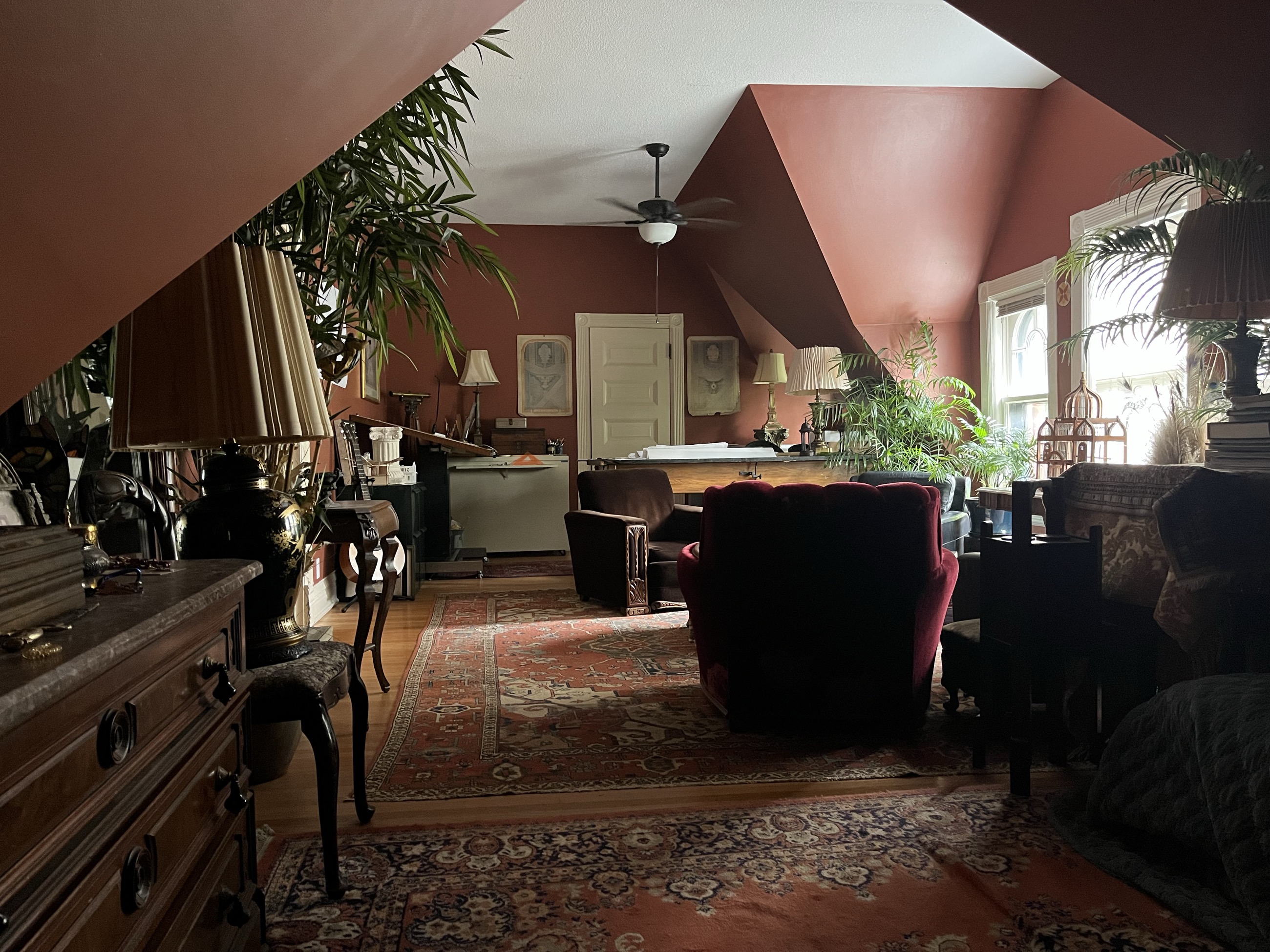 The studio interior — a long attic room with sloped rust-coloured ceilings, drafting table, worn rugs, velvet chairs and tall plants against the windows.