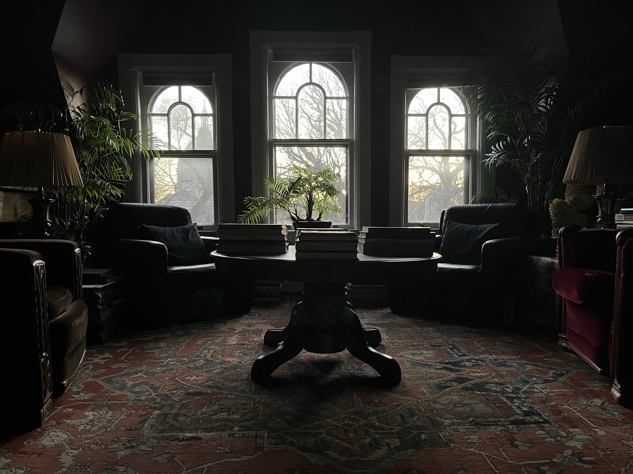 The studio at dusk — three arched windows above a round table stacked with books, lamps and palms in the dark corners of the room.